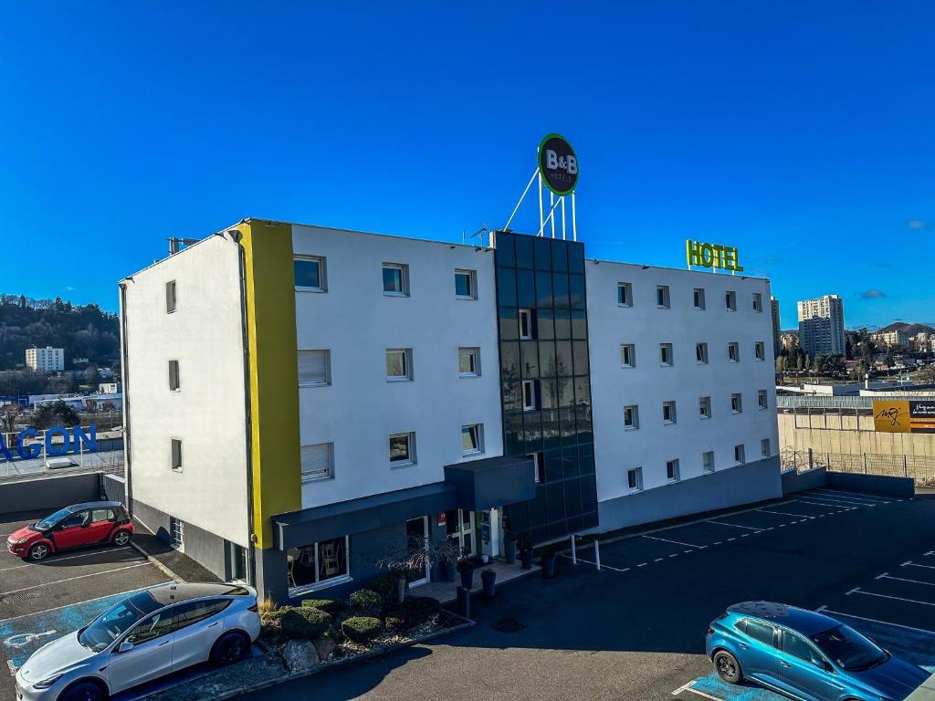 un bâtiment blanc avec des voitures garées dans un parking dans l'établissement B&B HOTEL Saint-Etienne Monthieu, à Saint-Étienne