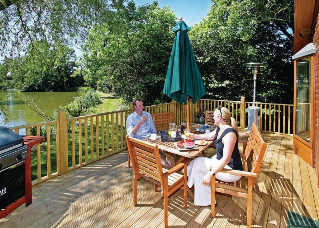 a man and woman sitting at a table on a deck with an umbrella at Upton Lakes Lodges in Cullompton