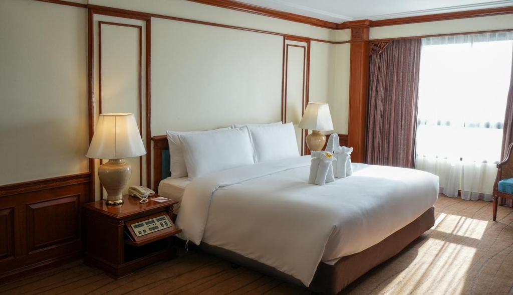 A large white bed is positioned against paneled walls beside a sunlit window.