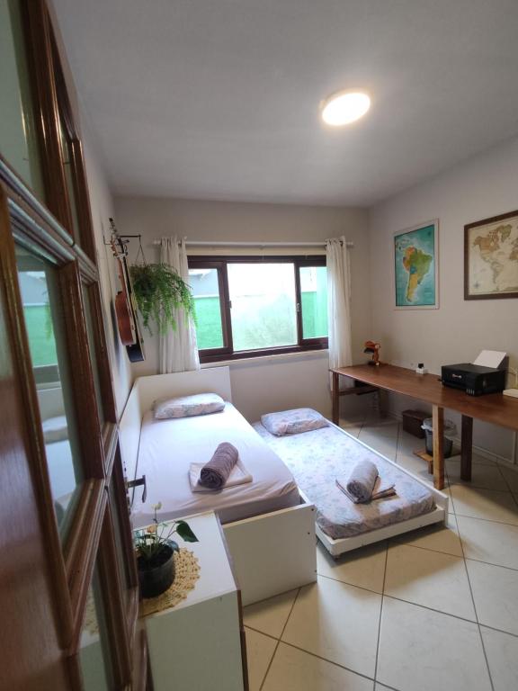 two beds in a room with a desk and a window at Quarto Felicidade Cabeçudas in Itajaí