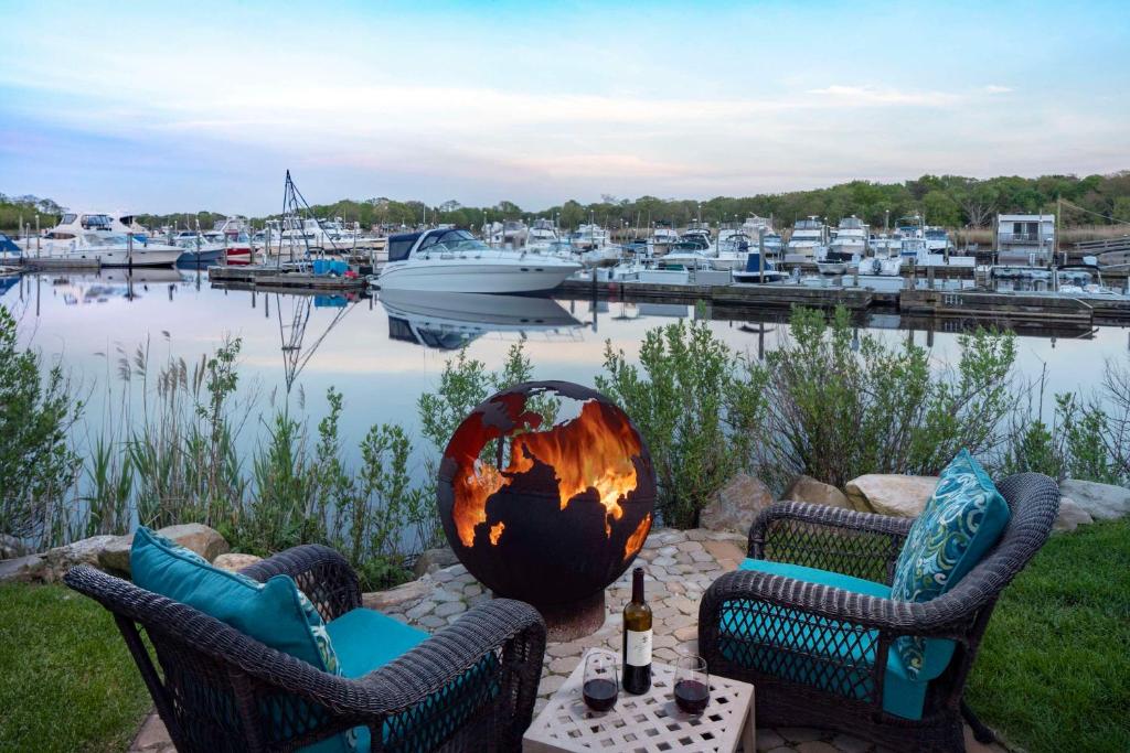 A photo of the grounds at Hyatt Place Long Island East End, featuring two large outdoor armchairs in turquoise blue, huddled around a large metal sculpture of a globe that doubles as a fire-pit, overlooking the water and the Marina with boats moored close by.