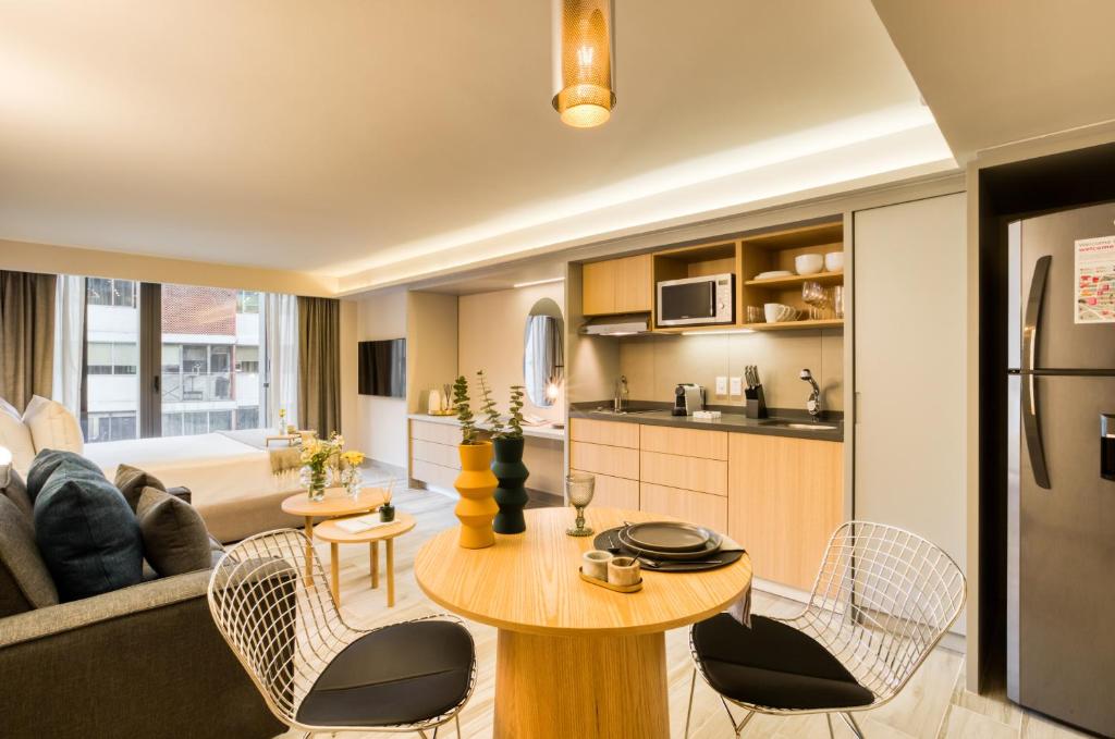 An open-plan studio combines a lounge, dining table, and full kitchenette beside a bright window.