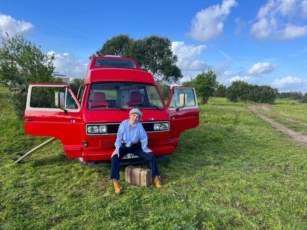 Uma mulher sentada numa mala em frente a uma carrinha vermelha. em Rent a Blue Classics' s Campervan for your Road trip in Portimao -VOLKSWAGEN T3 em Portimão