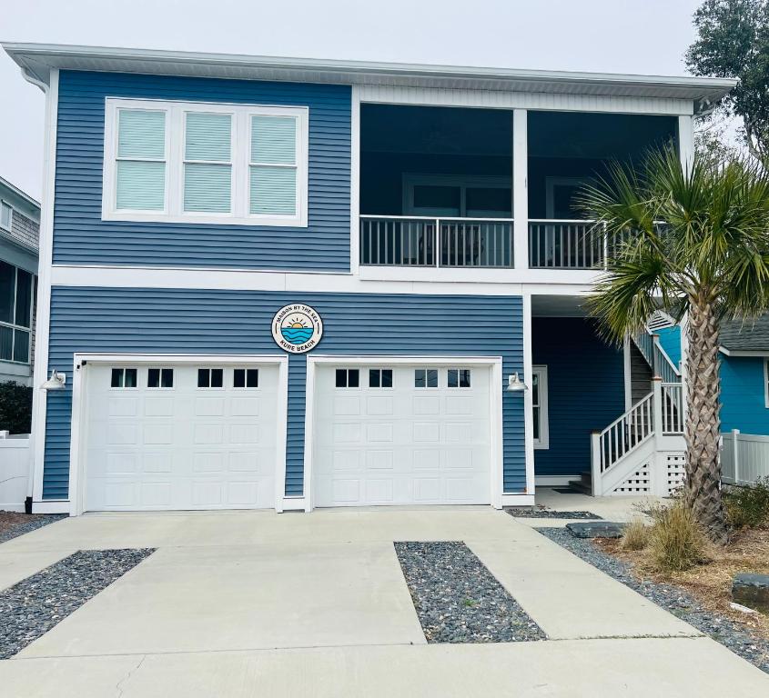 a blue house with white garage doors and a palm tree at Maison by The Sea at Kure Beach in Kure Beach