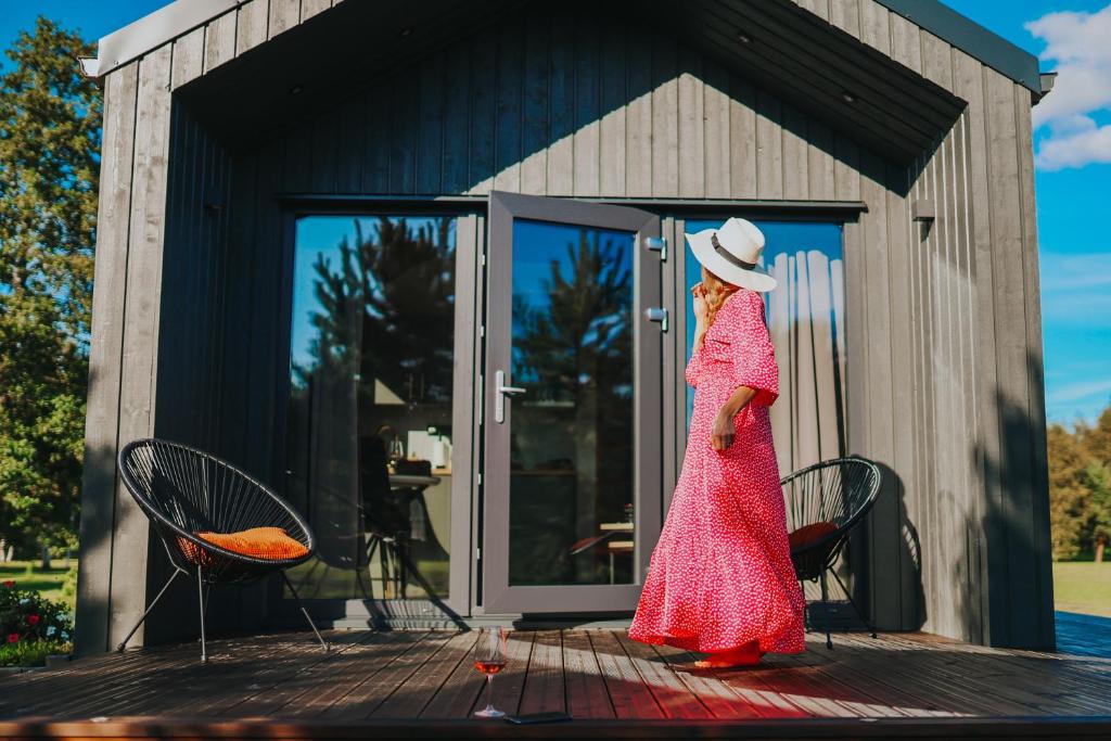 a woman in a red dress and hat standing on a deck at Atpūtas komplekss Pazust Priedēs - SUNSET house in Bernāti
