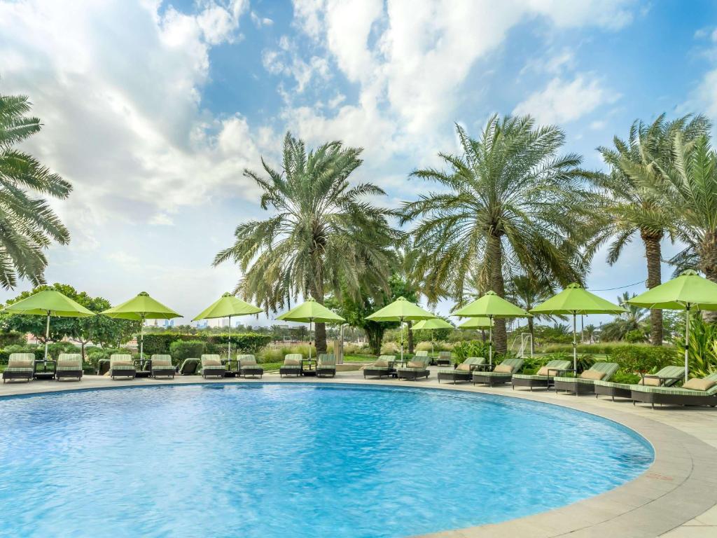 a pool with chairs and umbrellas and palm trees at Mövenpick Hotel Bahrain in Manama