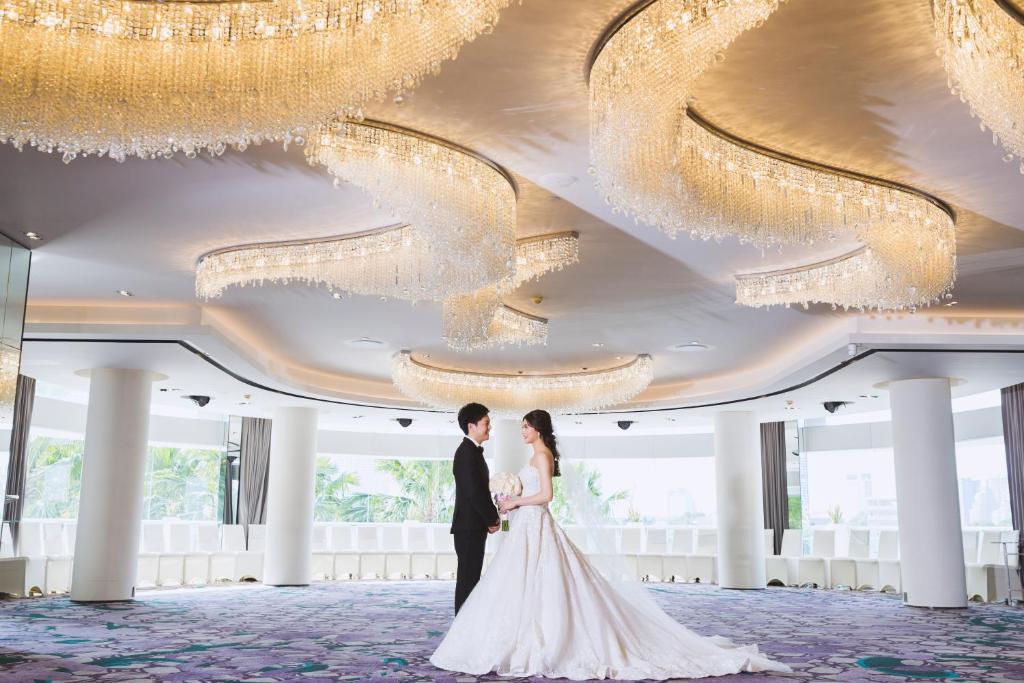 A bride and groom stand beneath chandeliers in a bright hotel lobby.