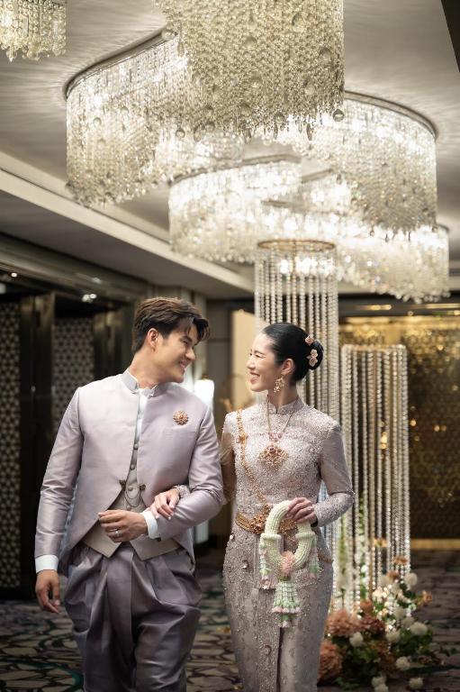 A formally dressed couple pose beneath a large chandelier in an ornate interior.