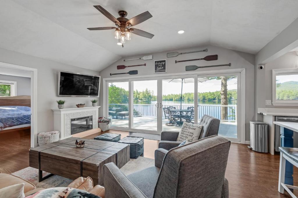 a living room with a fireplace and a ceiling fan at Waterfront Sun-Drenched Hermit Lake Retreat! in Sanbornton