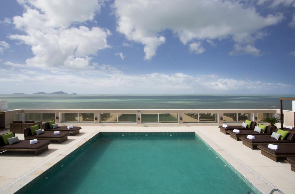 a swimming pool on top of a building with chairs and the ocean at Royal Atlântica Macaé Hotel in Macaé