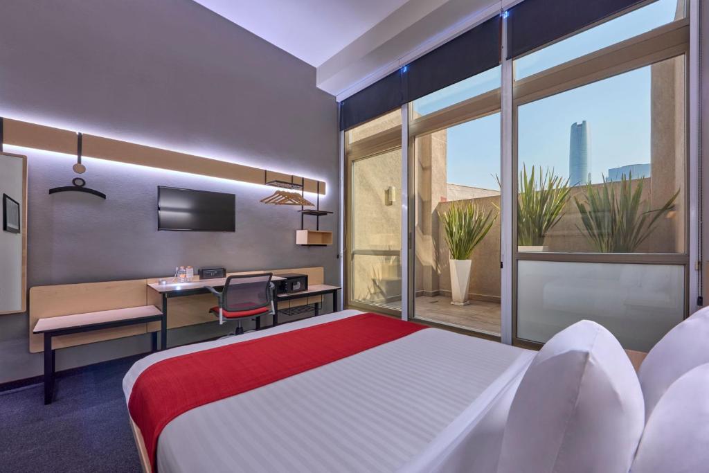 A modern room with a double bed, long work desk and glazed city-facing wall.