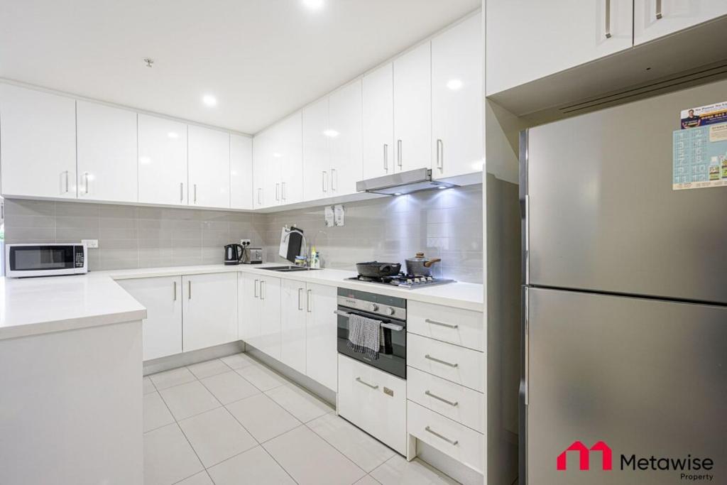 La cuisine est équipée de placards blancs et d'un réfrigérateur en acier inoxydable. dans l'établissement MetaWise Burwood Westfield Cozy 2Bed Free Parking VT108, à Sydney