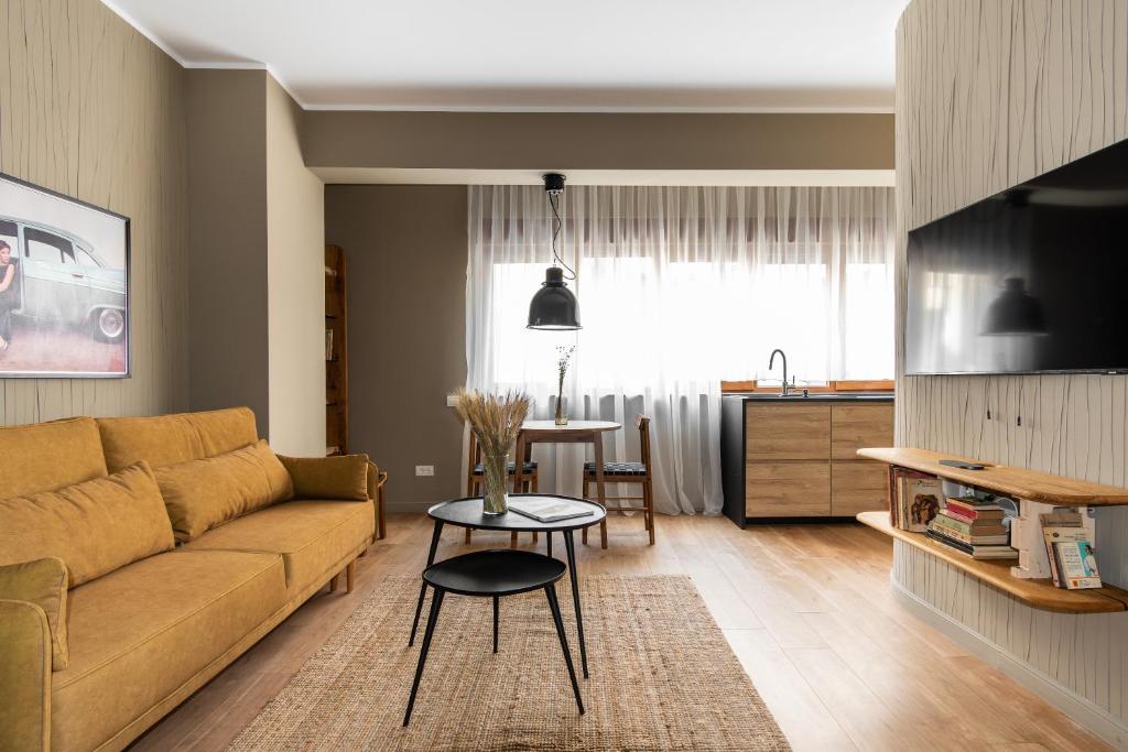 Open-plan studio with mustard sofa, wall-mounted TV, round dining table, sink in wood kitchenette and sheer curtains at Maison Bucarest Apartments by Olala Homes in Bucharest