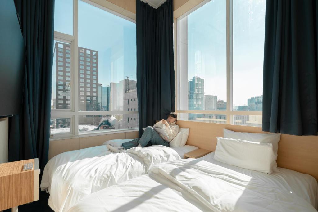 a man sitting on a bed in a room with windows at The Hyoo Sik ANNK Hong Dae Branch in Seoul