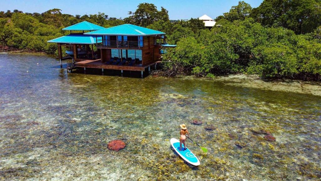 a woman standing on a surfboard in the water with a house at Bahia Coral Lodge in Bocas del Toro