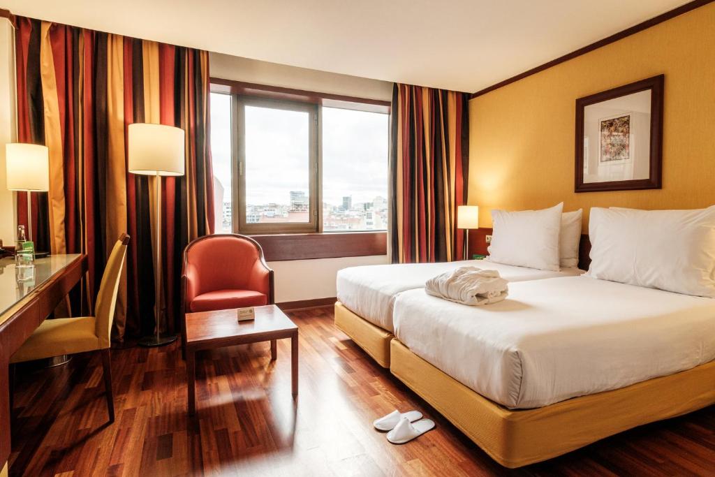 A warm-toned room with a large bed, compact desk, lounge chair and city-facing window.