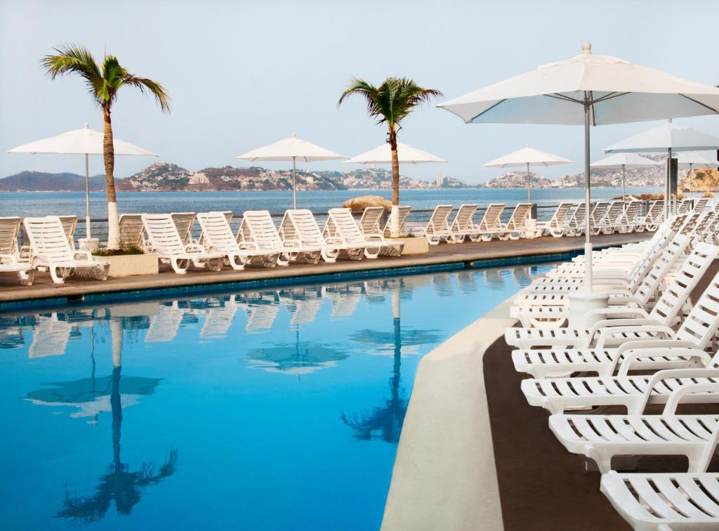 a swimming pool with white chairs and umbrellas at Calinda Beach Acapulco in Acapulco