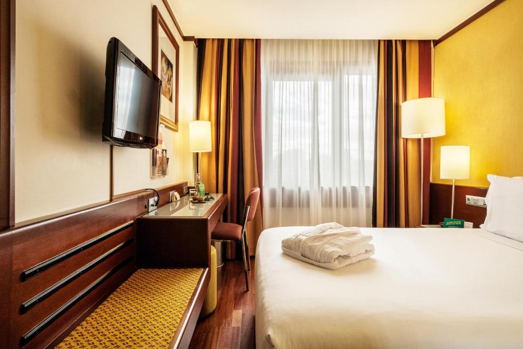 A guest room with a large bed, wall-mounted television, dresser and compact desk.