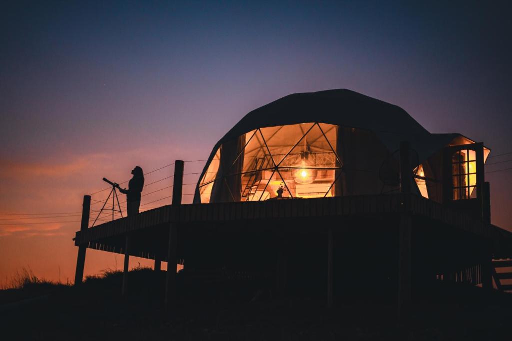 um homem parado em frente a uma cúpula ao pôr-do-sol em Glamping Atmosphera em Cambará