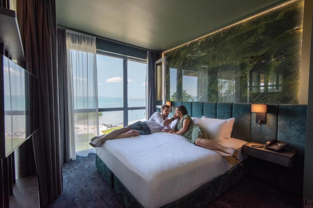 a man and woman sitting on a bed in a hotel room at Hotel Azur Premium in Siófok