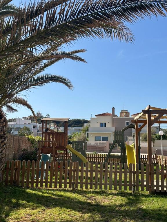 a wooden fence with a playground behind a palm tree at Nautica Hotel Apartments in Stavromenos