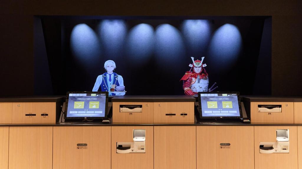 Henn na Hotel Kagoshima Tenmonkan في كاجوشيما: شاشة تلفزيون مع رجل و جهازين كمبيوتر محمول على مكتب