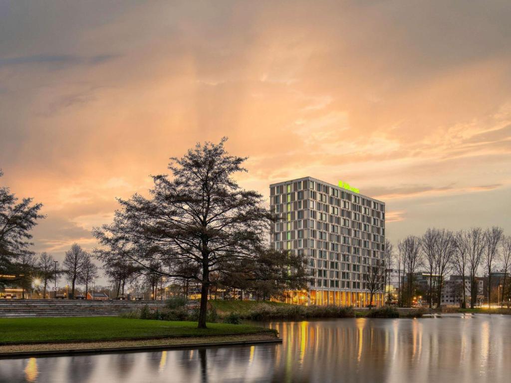 un edificio alto con un albero di fronte a un lago di ibis Styles Rotterdam Ahoy a Rotterdam