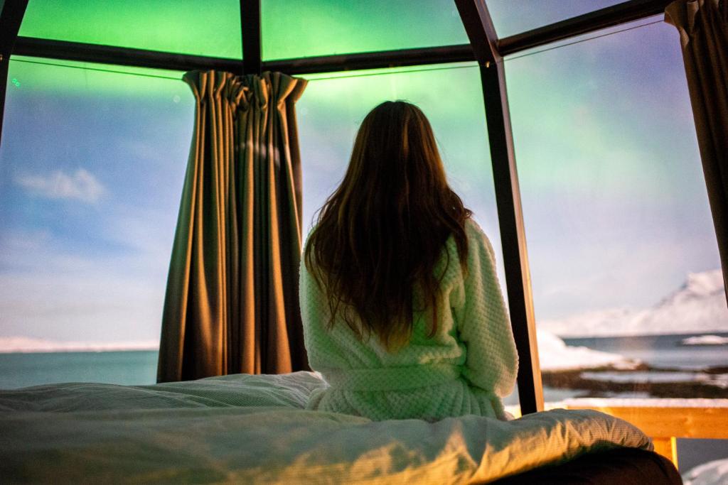 una mujer sentada en una cama mirando por la ventana en Aurora Hut by InukTravel en Nuuk