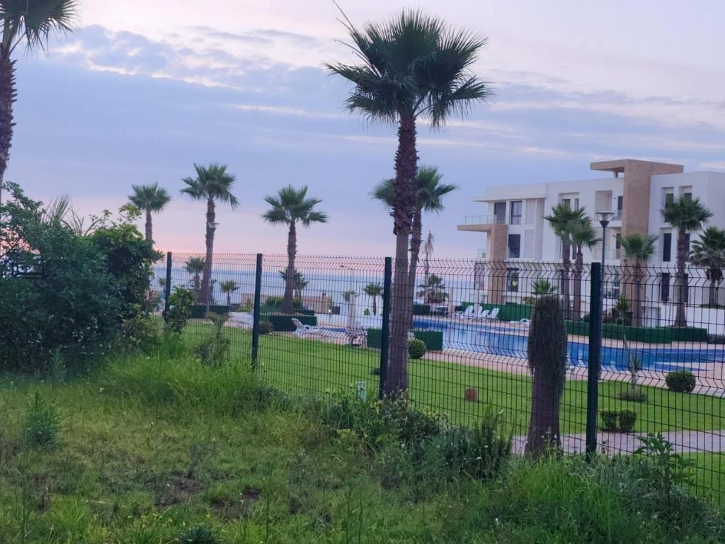 a fence with palm trees in front of a pool at Plage dès nations 2 bedroom apartment with backyard view in Sidi Bouqnadel