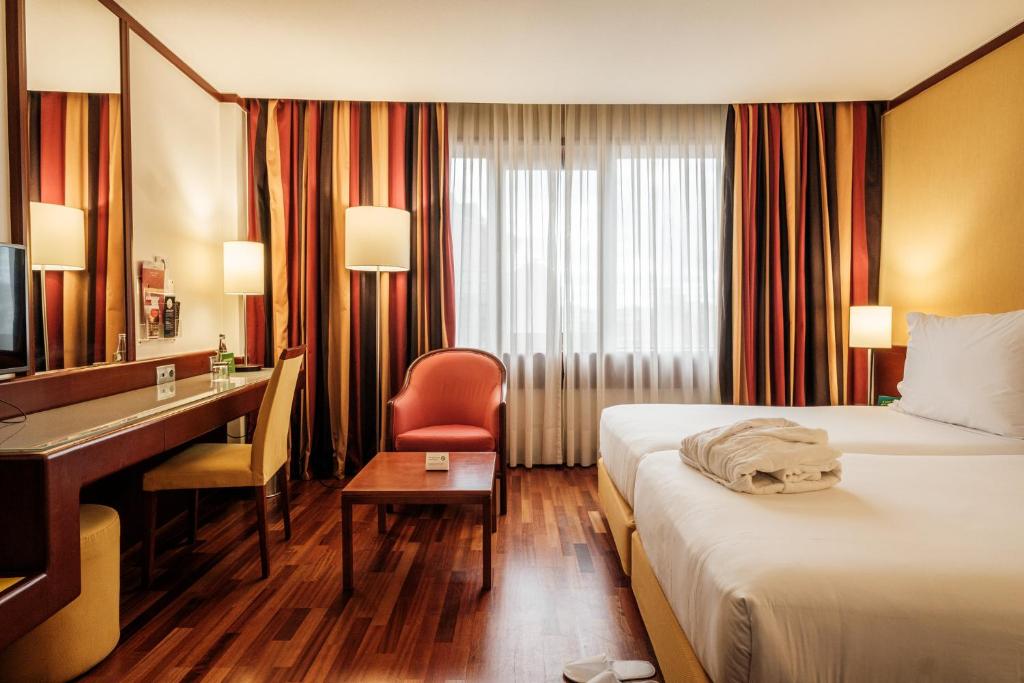 A guest room viewed from the desk side, with a bed, armchair and bright curtained window.