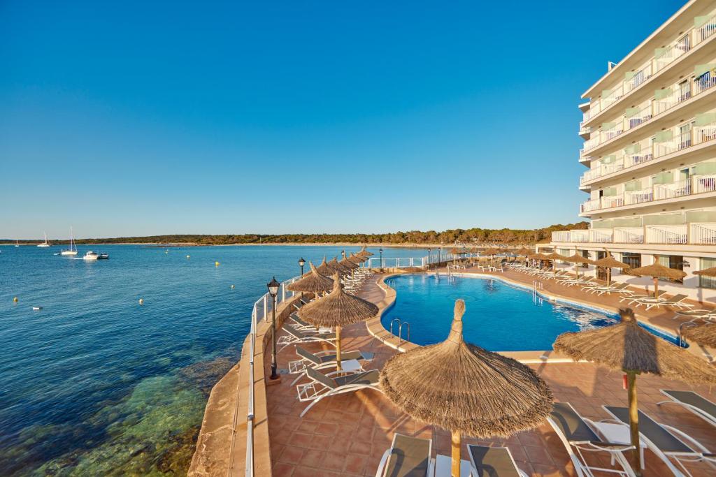 a resort with a swimming pool with straw umbrellas and the ocean at Universal Hotel Marqués in Colònia de Sant Jordi