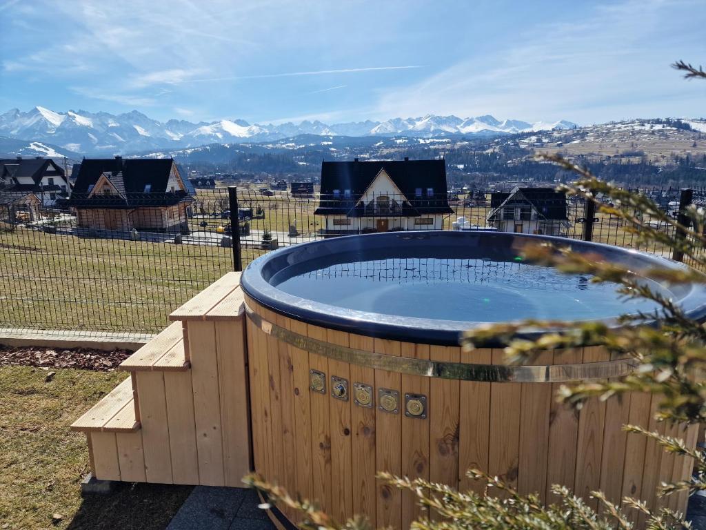 Wooden hot tub on lawn overlooking village rooftops and distant snowy mountains under clear blue sky.
