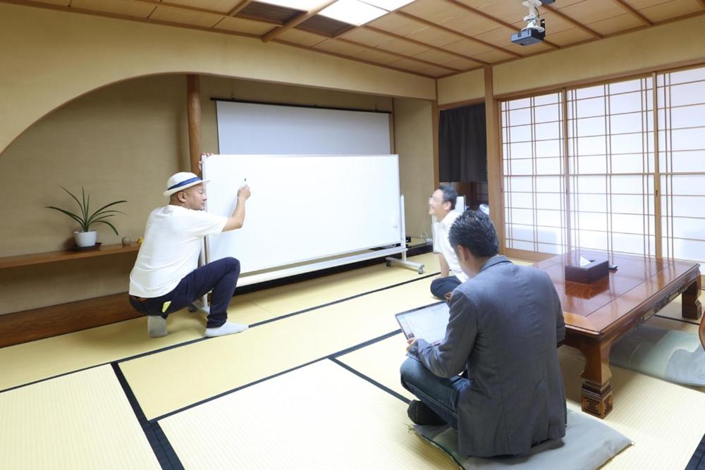 un gruppo di persone sedute in una stanza con una presentazione di 熱海の隠れ里 ad Atami