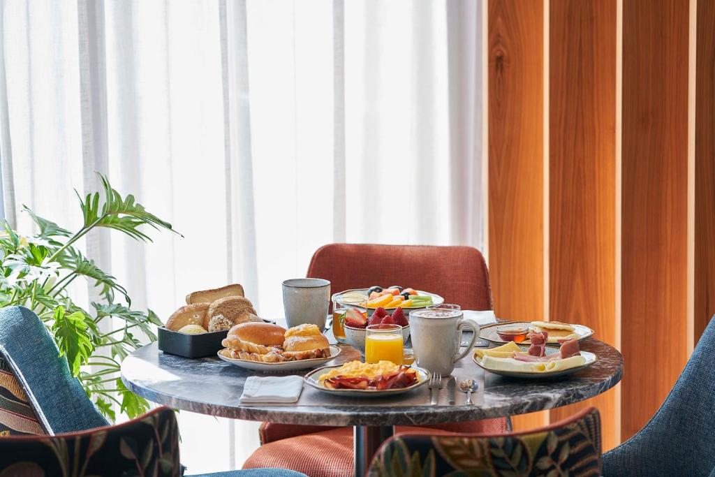 A breakfast table displays pastries, bread, fruit, yogurt and coffee beside a bright curtained window.