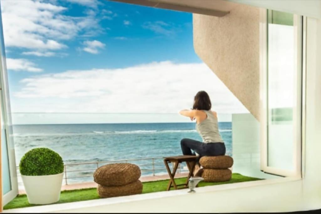 a woman sitting on a stool looking out a window at the ocean at OceanSound White luminoso y tranquilo in Las Palmas de Gran Canaria