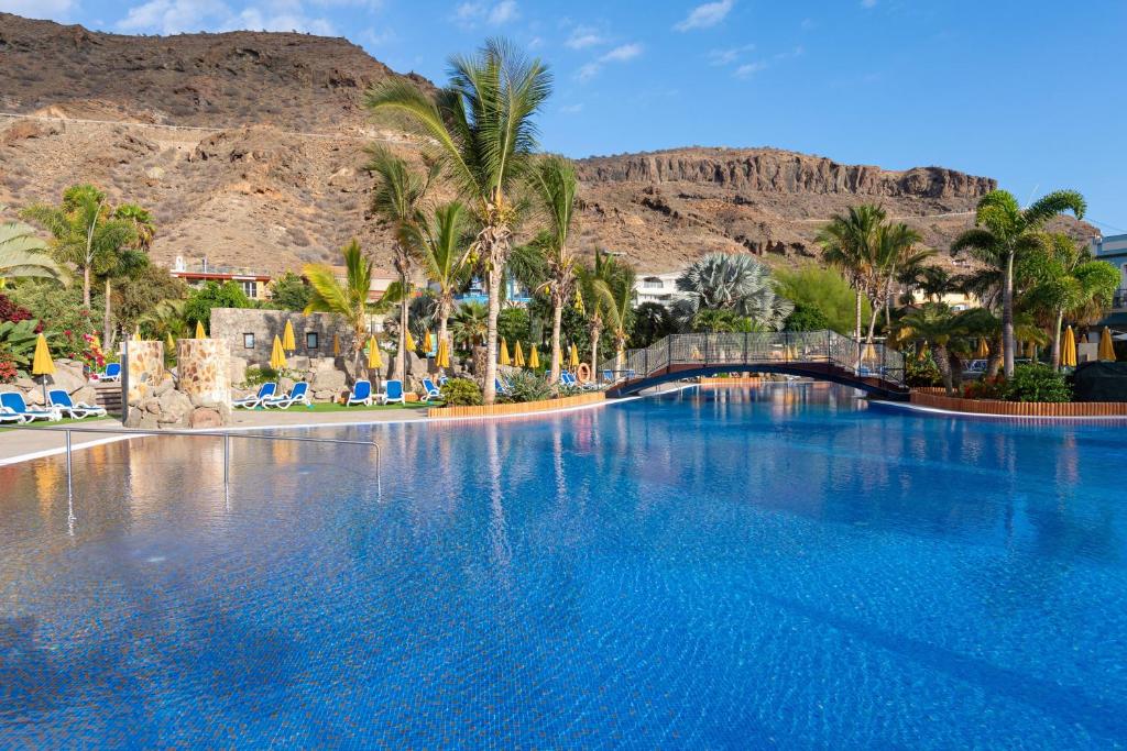 a large swimming pool with palm trees and mountains at Hotel Cordial Mogán Playa in Puerto de Mogán