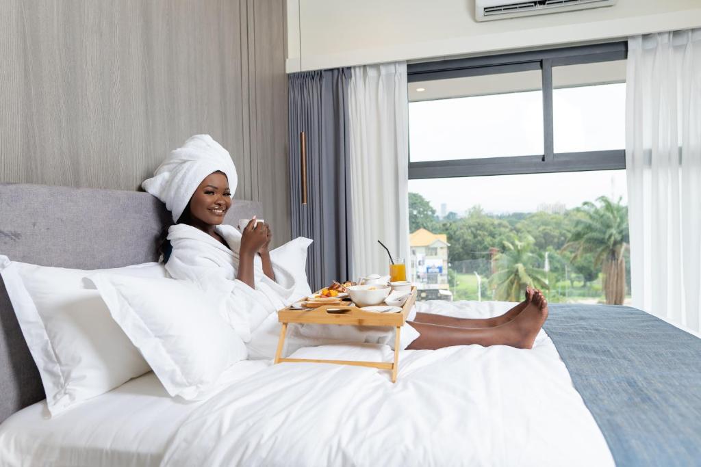 Eine Frau sitzt mit einem Tablett voller Essen auf einem Bett. in der Unterkunft Lux Inn Hotel in Kinshasa