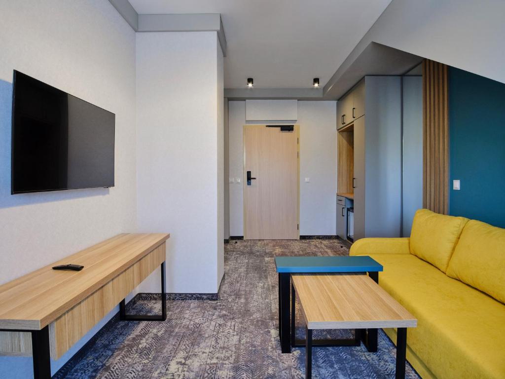 Hotel living area with wall-mounted TV, wooden desk, yellow sofa, nesting coffee tables.