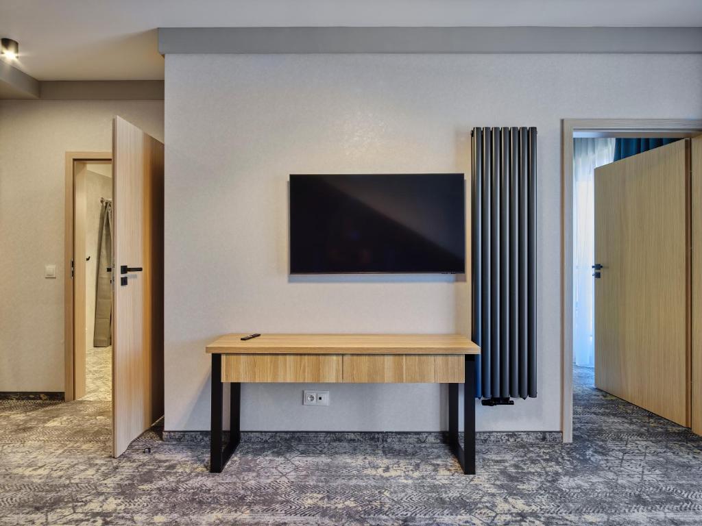 Wall-mounted flat-screen TV above wooden desk in hotel room corridor, open doors and radiator visible.