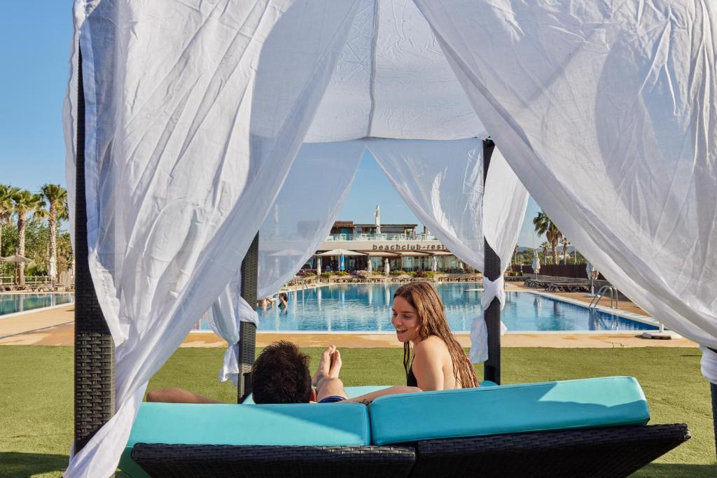Dos mujeres están sentadas en un sofá debajo de una tienda de campaña cerca de una piscina. en TAIGA Delta de l'Ebre, en L'Ampolla