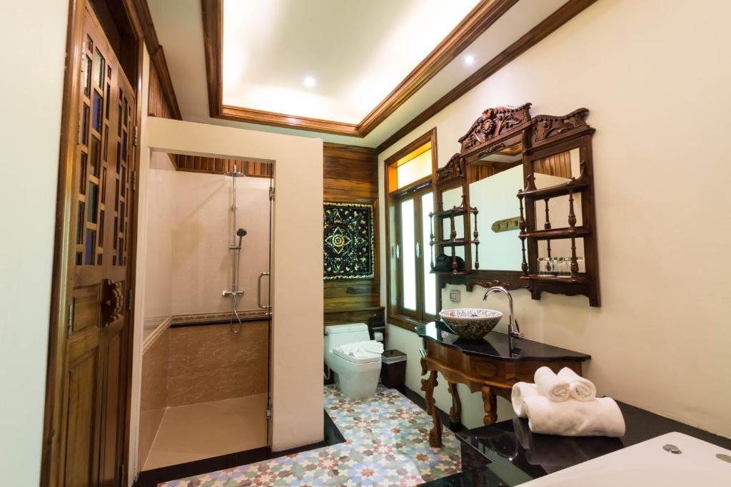 An ornate bathroom combines a glass-enclosed shower, carved vanity, toilet, and patterned tile floor.