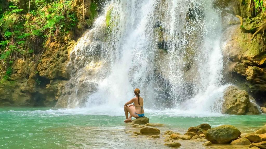 Una donna seduta sulle rocce di fronte a una cascata di Beach Apartment on Samana Bay with Pool, AC, Fast Wi-Fi, Walk to Beach & River, Whale Watching, Near Salto El Limón a Santa Bárbara de Samaná