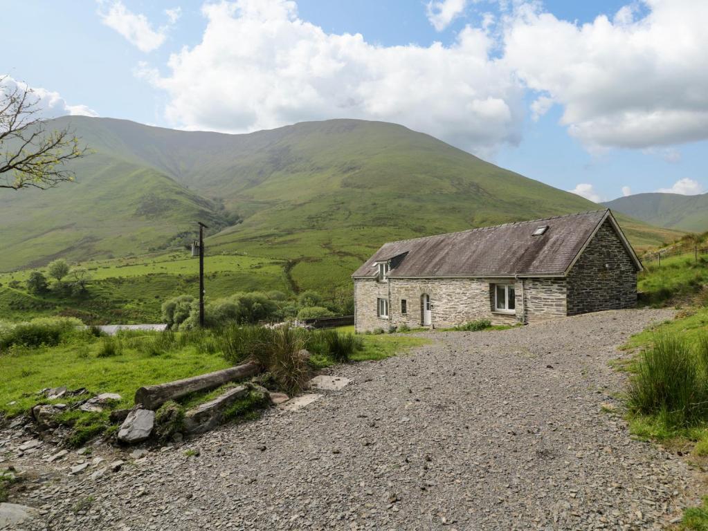Una antigua cabaña de piedra en un camino de grava con montañas al fondo en Pennant Tigi Uchaf, en Dolgellau