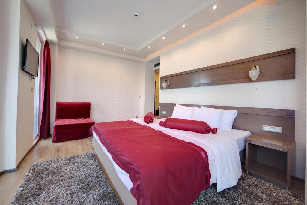 Modern hotel room with king bed and red runner, wall-mounted TV, bedside lights, chaise lounge and textured accent wall at City Palace Hotel in Ohrid