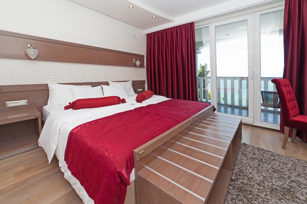Sea-view room with king bed, red throw and curtains, wooden headboard, bedside lamps and balcony with chair at City Palace Hotel in Ohrid