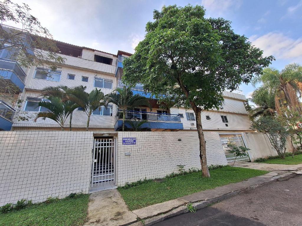 Un edificio blanco con un árbol delante. en Quarto em Apto confortável 10 min da praia do Recreio RJ, en Río de Janeiro