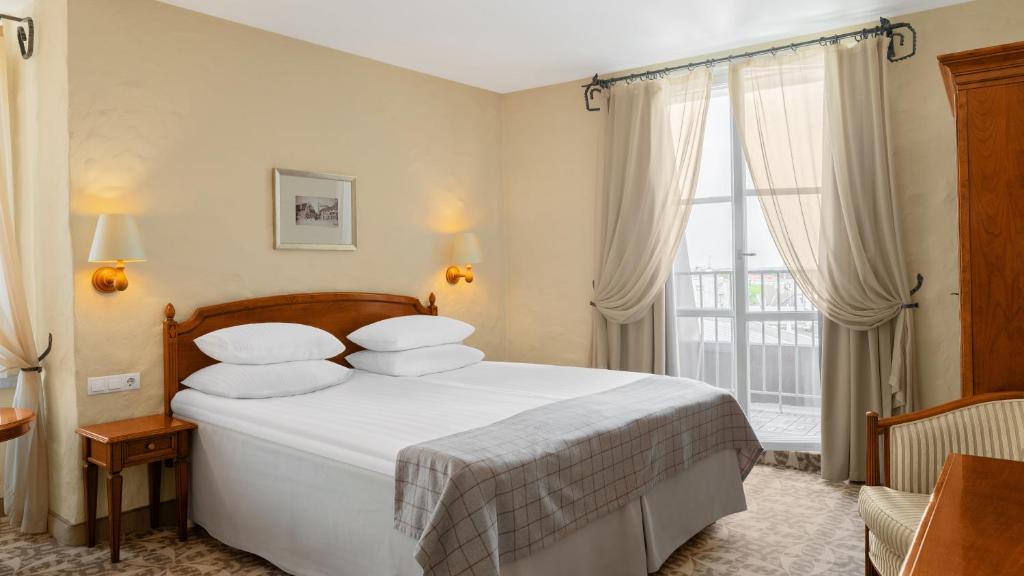 Double bed with wooden headboard, wall sconce and bedside lamp, striped chair, and balcony doors with sheer curtains at Rixwell Collection Savoy Boutique Hotel in Tallinn