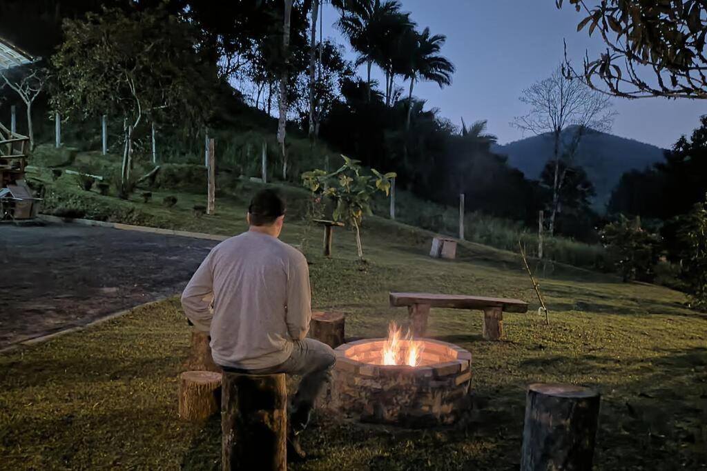a man sitting on a bench next to a fire at Casa de campo rústica 21km vila germânica Bnu in Blumenau