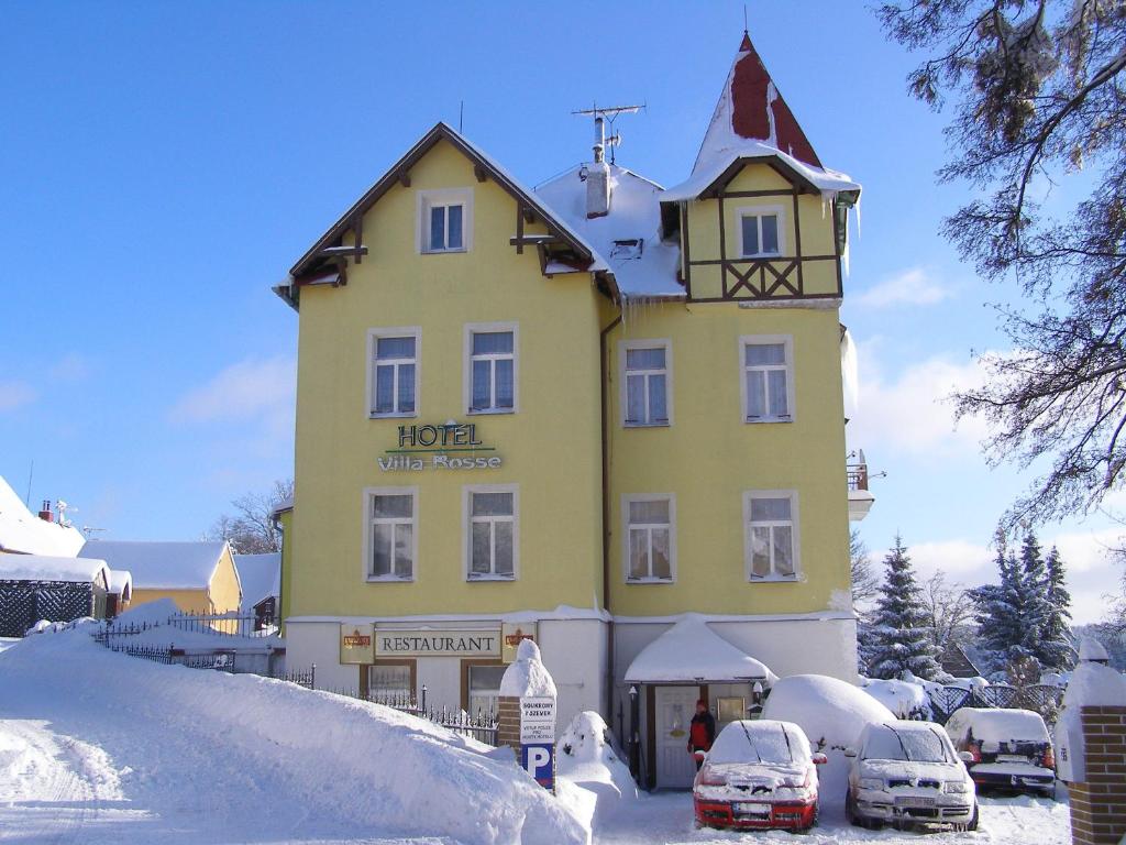 un edificio amarillo en la nieve con coches aparcados delante en Penzion Villa Rosse, en Abertamy