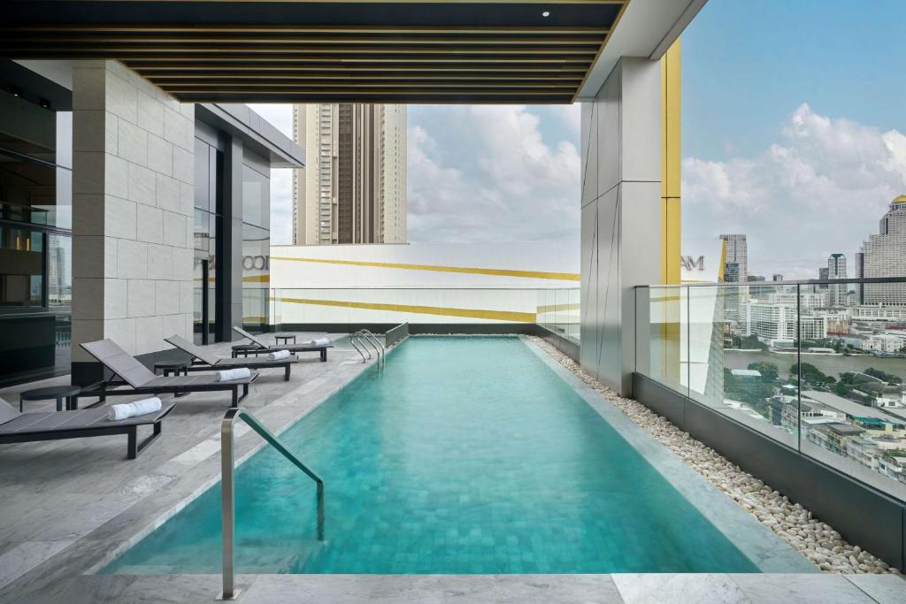 A long rooftop pool runs beside loungers and a glass railing overlooking Bangkok.